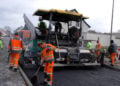 Straßenfertiger baut eine neue Asphaltdecke ein, Bauarbeiter in orangefarbener Warnkleidung ziehen den Belag ab