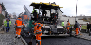 Straßenfertiger baut eine neue Asphaltdecke ein, Bauarbeiter in orangefarbener Warnkleidung ziehen den Belag ab