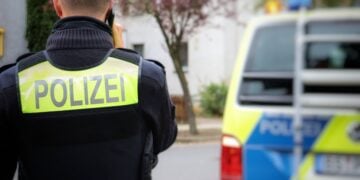Polizist mit Funkgerät vor einem Wohnhaus, daneben ein Streifenwagen