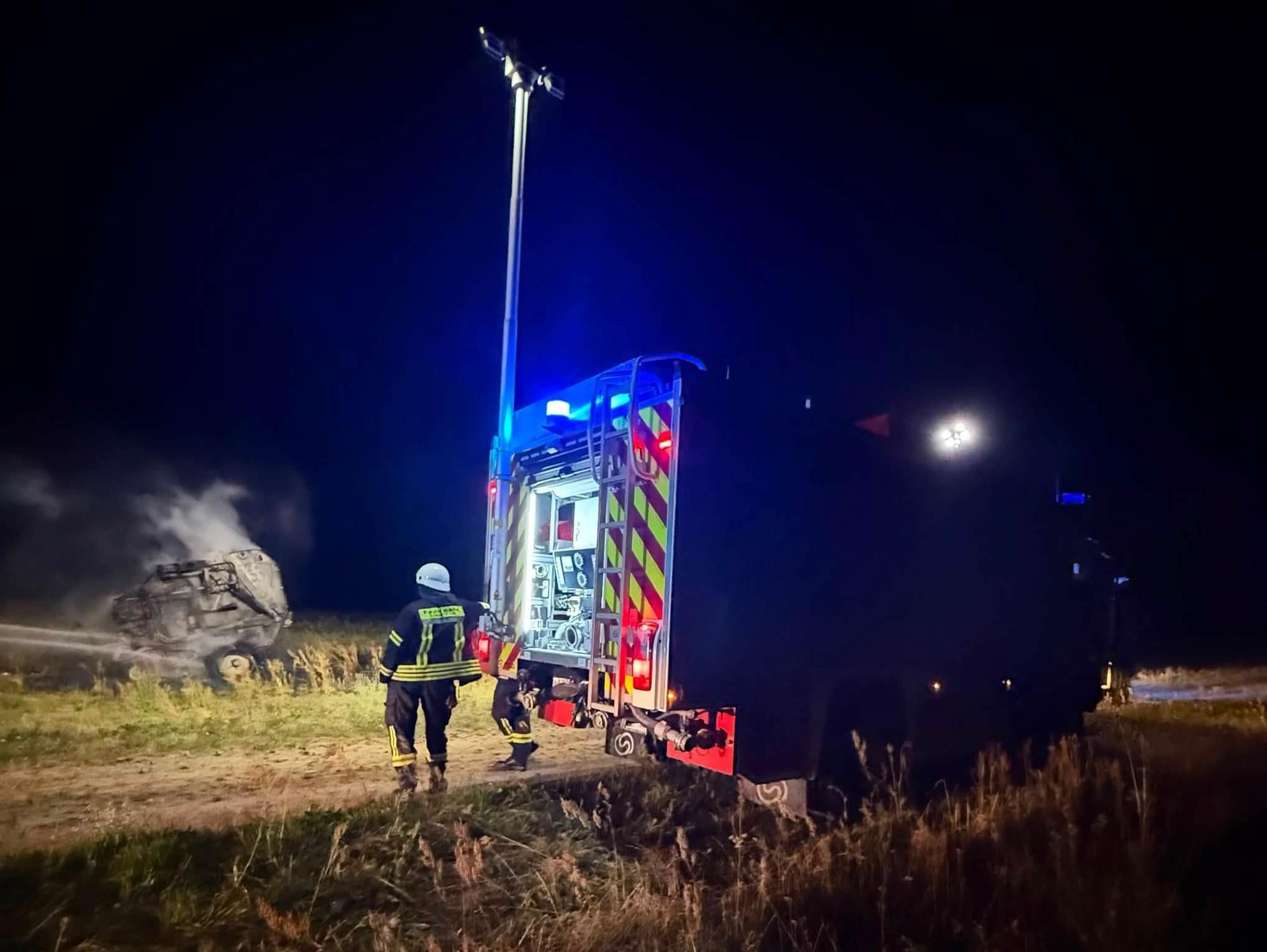 Löschfahrzeug mit ausgefahrenem Lichtmast steht nachts an einem Feldweg, im Hintergrund raucht die Brandstelle