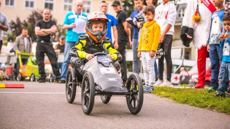 Ein Kind mit Helm steuert eine Seifenkiste an Zuschauern vorbei