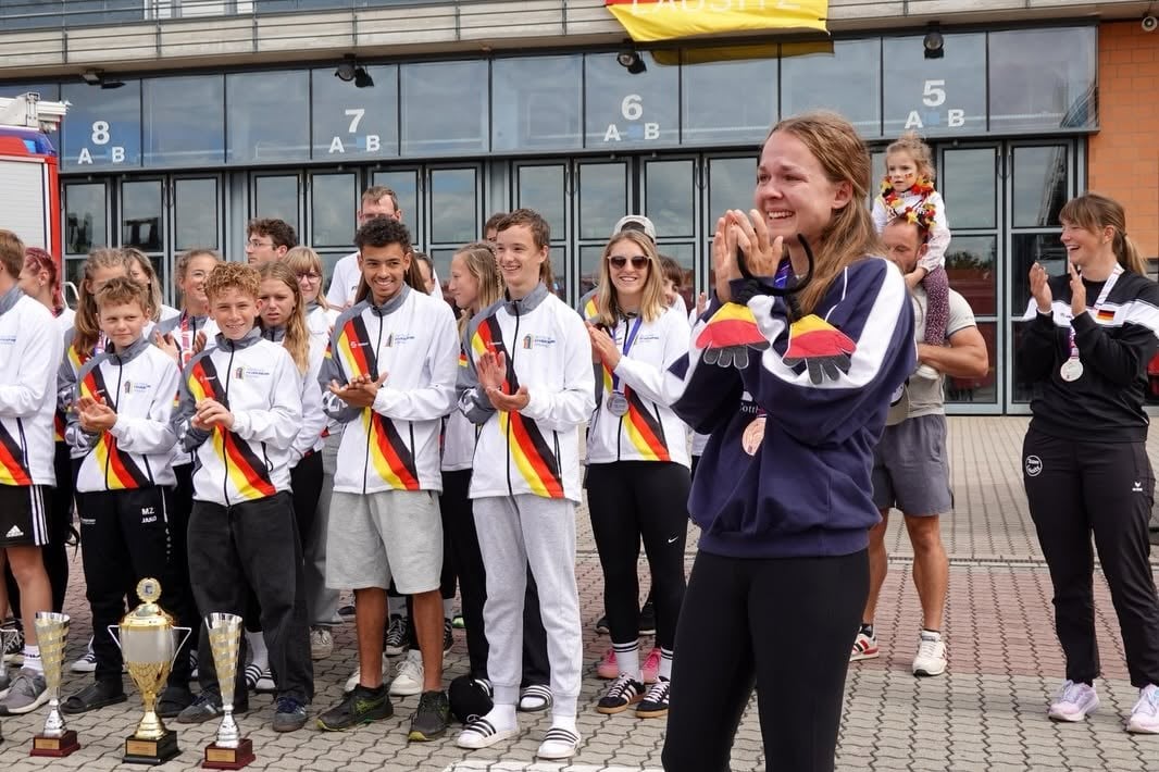 Sportlerin zeigt lachend ihre Medaille, dahinter applaudiert das Team