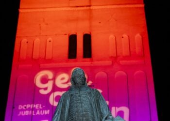 Illuminierte Kirchturmfassade mit dem Denkmal Paul Gerhardts davor