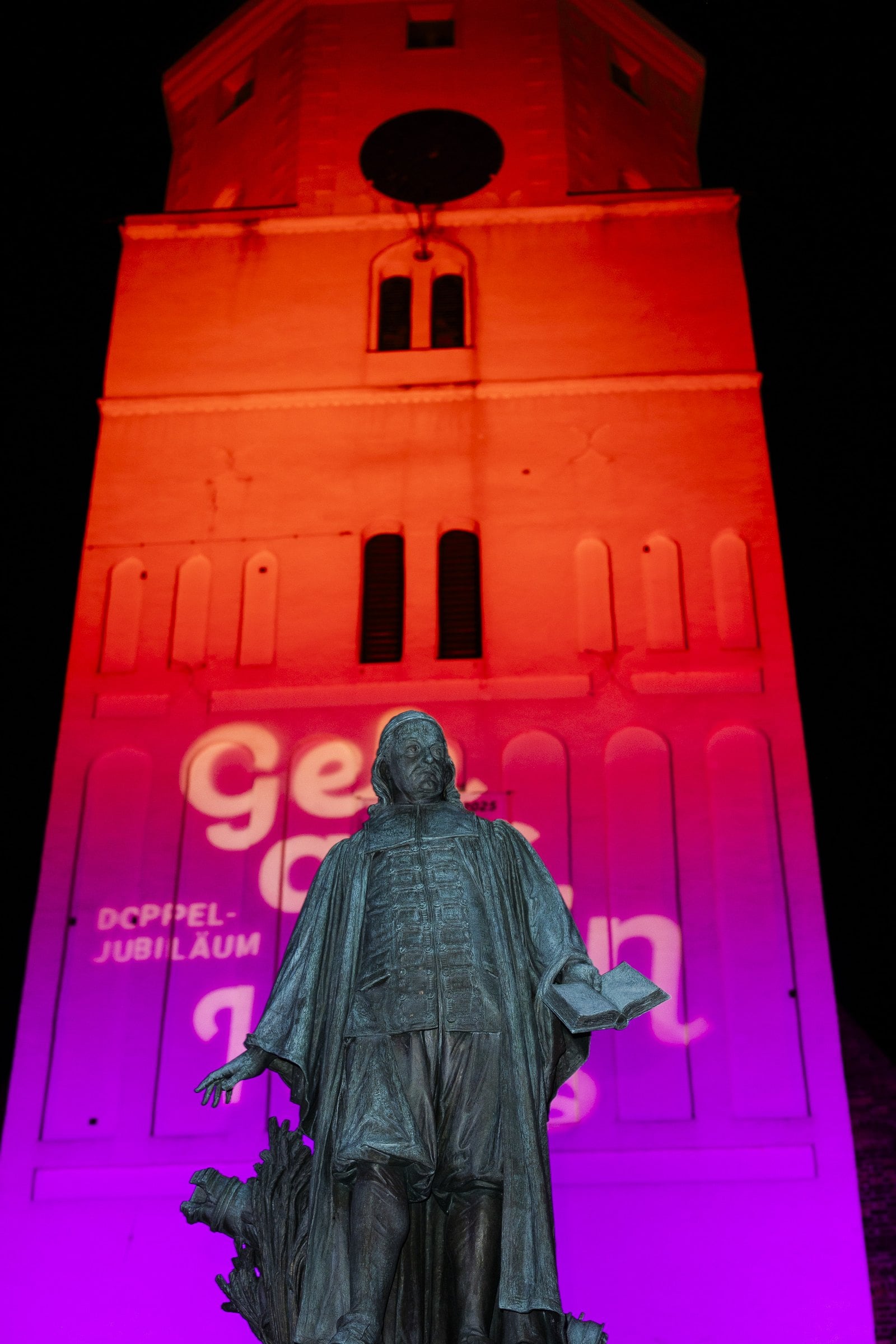 Illuminierte Kirchturmfassade mit dem Denkmal Paul Gerhardts davor