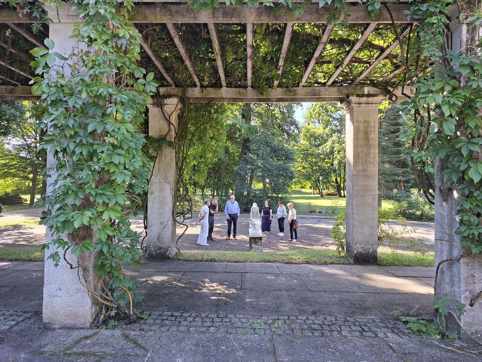 Blick durch die begrünte Pergola auf eine Gruppe von Menschen um das Tonmodell der Wassernixe