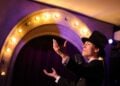 Ein Darsteller mit Zylinder singt in ein Standmikrofon vor einem beleuchteten Bühnenportal