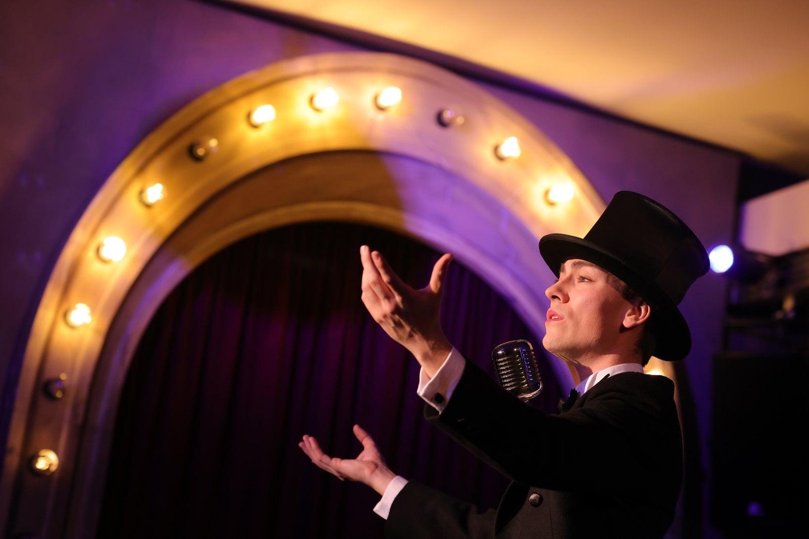 Ein Darsteller mit Zylinder singt in ein Standmikrofon vor einem beleuchteten Bühnenportal