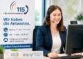 Mitarbeiterin mit Headset am Arbeitsplatz neben einem Aufsteller zur Behördennummer 115