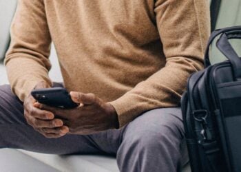 Sitzende Person mit Smartphone, daneben eine schwarze Laptoptasche
