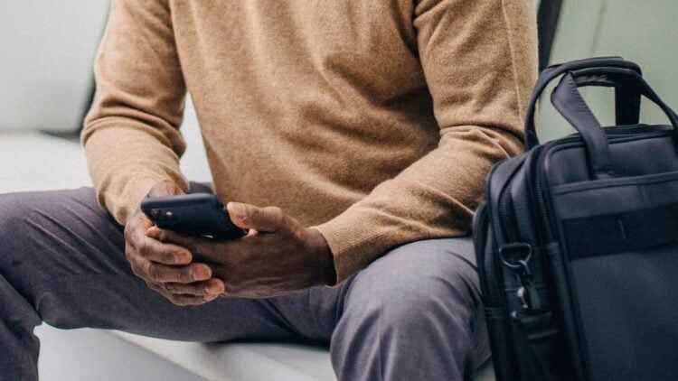Sitzende Person mit Smartphone, daneben eine schwarze Laptoptasche