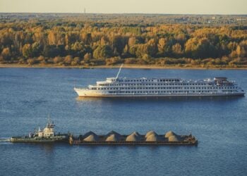 Weisses Flusskreuzfahrtschiff faehrt auf einem breiten Fluss, dahinter herbstlicher Wald