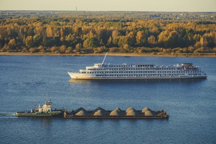 Weisses Flusskreuzfahrtschiff faehrt auf einem breiten Fluss, dahinter herbstlicher Wald
