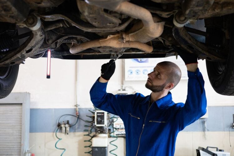 Mechaniker im blauen Overall arbeitet unter einem aufgebockten Auto an der Abgasanlage
