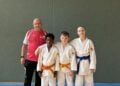 Drei junge Judoka in weißen Anzügen stehen neben ihrem Trainer in einer Sporthalle