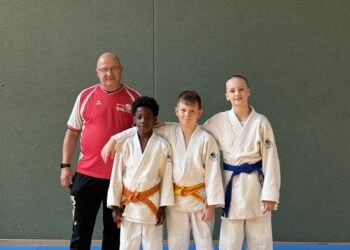 Drei junge Judoka in weißen Anzügen stehen neben ihrem Trainer in einer Sporthalle