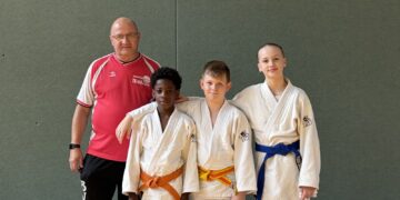 Drei junge Judoka in weißen Anzügen stehen neben ihrem Trainer in einer Sporthalle