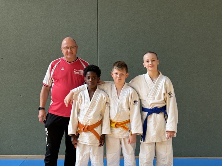 Drei junge Judoka in weißen Anzügen stehen neben ihrem Trainer in einer Sporthalle