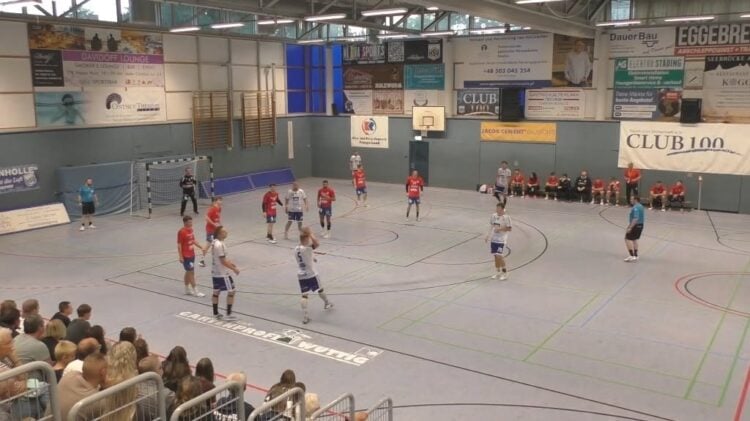 Handballspiel in einer Sporthalle, Spieler in roten und weißen Trikots im Angriff