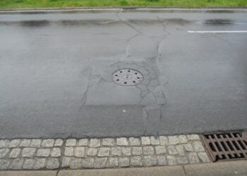 Rissige Asphaltdecke um einen Schachtdeckel, daneben eine Natursteinpflasterrinne