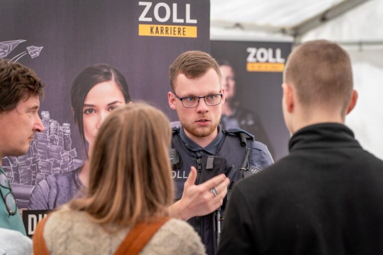 Ein Zollbeamter spricht mit zwei Besuchern vor einer Messewand