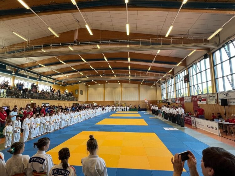 Junge Judoka in weißen Anzügen stehen zur Eröffnung um die Wettkampfmatte in einer Sporthalle