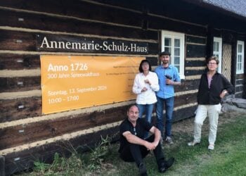 Vier Menschen stehen vor einem Blockbohlenhaus mit einem Plakat zum 300-jährigen Jubiläum