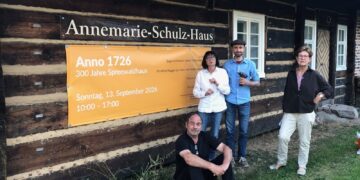 Vier Menschen stehen vor einem Blockbohlenhaus mit einem Plakat zum 300-jährigen Jubiläum