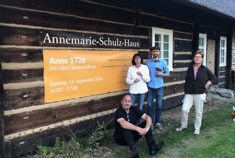 Vier Menschen stehen vor einem Blockbohlenhaus mit einem Plakat zum 300-jährigen Jubiläum