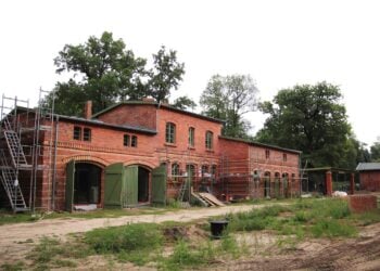 Langgestreckter Backsteinbau mit grünen Toren und Baugerüst, davor unbefestigter Boden