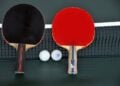 Zwei Tischtennisschläger in Rot und Schwarz mit zwei Bällen am Netz einer grünen Platte