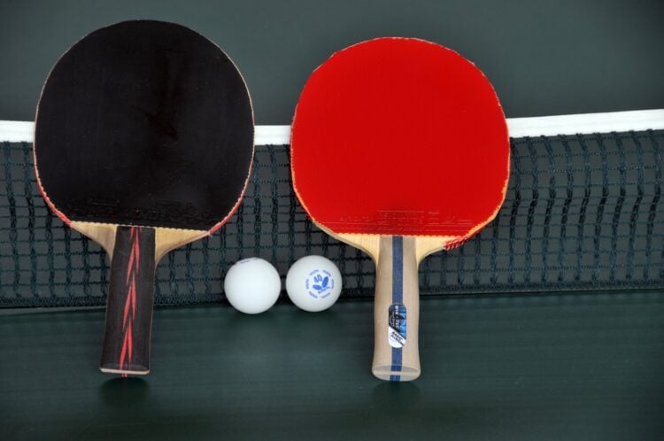 Zwei Tischtennisschläger in Rot und Schwarz mit zwei Bällen am Netz einer grünen Platte