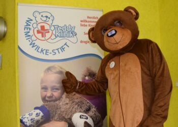 Bär im Kostüm steht neben dem Banner der Teddy-Klinik