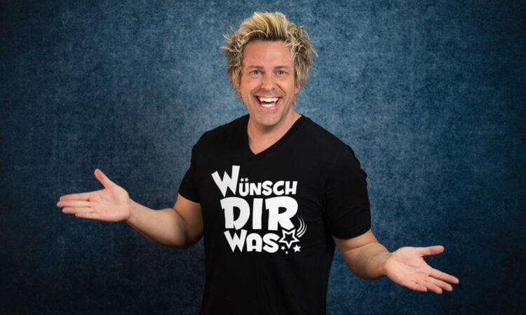 Sascha Grammel im schwarzen T-Shirt mit der Aufschrift Wünsch Dir Was, lachend mit ausgebreiteten Armen