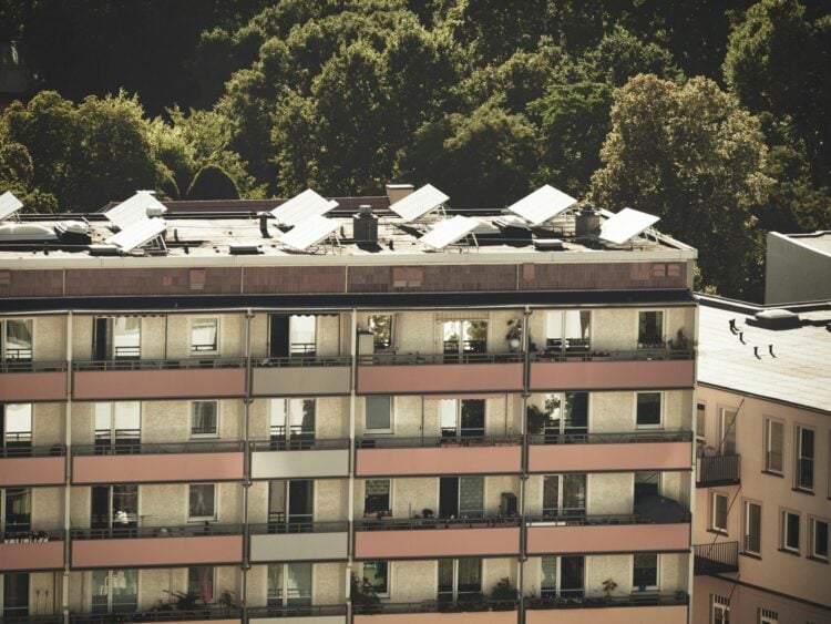Mehrgeschossiger Wohnblock mit Balkonen, auf dem Flachdach stehen Solarkollektoren, dahinter Baumkronen