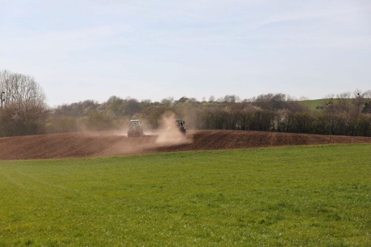 Zwei Traktoren bearbeiten ein Feld und wirbeln Staub auf, im Vordergrund eine grüne Wiese