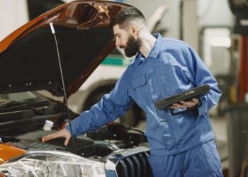 Kfz-Mechaniker in blauer Arbeitskleidung steht mit einem Diagnosetablett vor dem geöffneten Motorraum eines Autos in einer Werkstatt