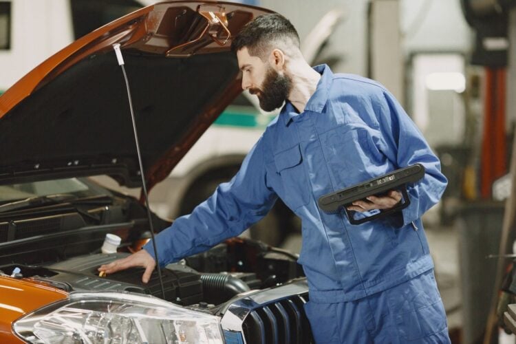 Kfz-Mechaniker in blauer Arbeitskleidung steht mit einem Diagnosetablett vor dem geöffneten Motorraum eines Autos in einer Werkstatt