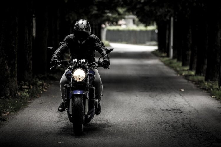 Motorradfahrer mit Integralhelm und Motorradjacke fährt auf einer von Bäumen gesäumten Straße frontal auf die Kamera zu