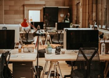 Mehrere Schreibtische in einem offenen Büro, darauf Bildschirme, Schreibtischlampen und persönliche Gegenstände