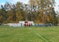 Zwei Fußballmannschaften in Weiß und Blau-Gelb stehen vor dem Anpfiff an der Mittellinie, dahinter Bandenwerbung und Herbstbäume