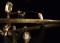 Puppenspieler mit weiß geschminktem Gesicht führt eine geschnitzte Stabpuppe über einen Holzbalken