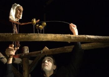Puppenspieler mit weiß geschminktem Gesicht führt eine geschnitzte Stabpuppe über einen Holzbalken