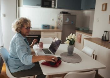 Ältere Frau mit Brille sitzt an einem Küchentisch und arbeitet am Laptop
