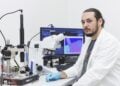 Aleksandar Matkovic im weißen Laborkittel an einem Mikroskop, im Hintergrund ein Monitor