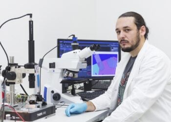 Aleksandar Matkovic im weißen Laborkittel an einem Mikroskop, im Hintergrund ein Monitor