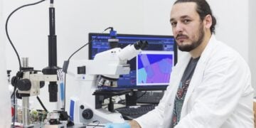 Aleksandar Matkovic im weißen Laborkittel an einem Mikroskop, im Hintergrund ein Monitor