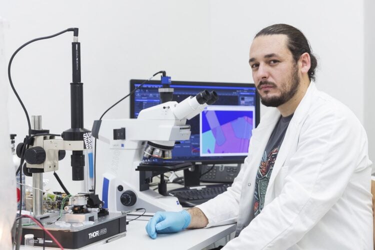 Aleksandar Matkovic im weißen Laborkittel an einem Mikroskop, im Hintergrund ein Monitor