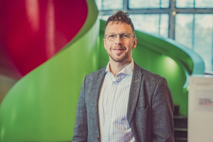André Wendler mit Brille und Sakko vor der roten und grünen Wendeltreppe im IKMZ in Cottbus