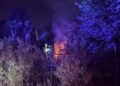 Zwei Feuerwehrleute mit Helmen hinter einem Maschendrahtzaun, daneben brennende Holzpaletten zwischen Büschen bei Nacht