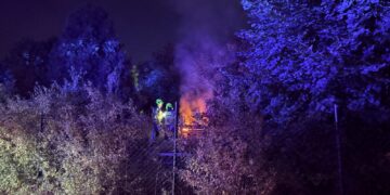 Zwei Feuerwehrleute mit Helmen hinter einem Maschendrahtzaun, daneben brennende Holzpaletten zwischen Büschen bei Nacht